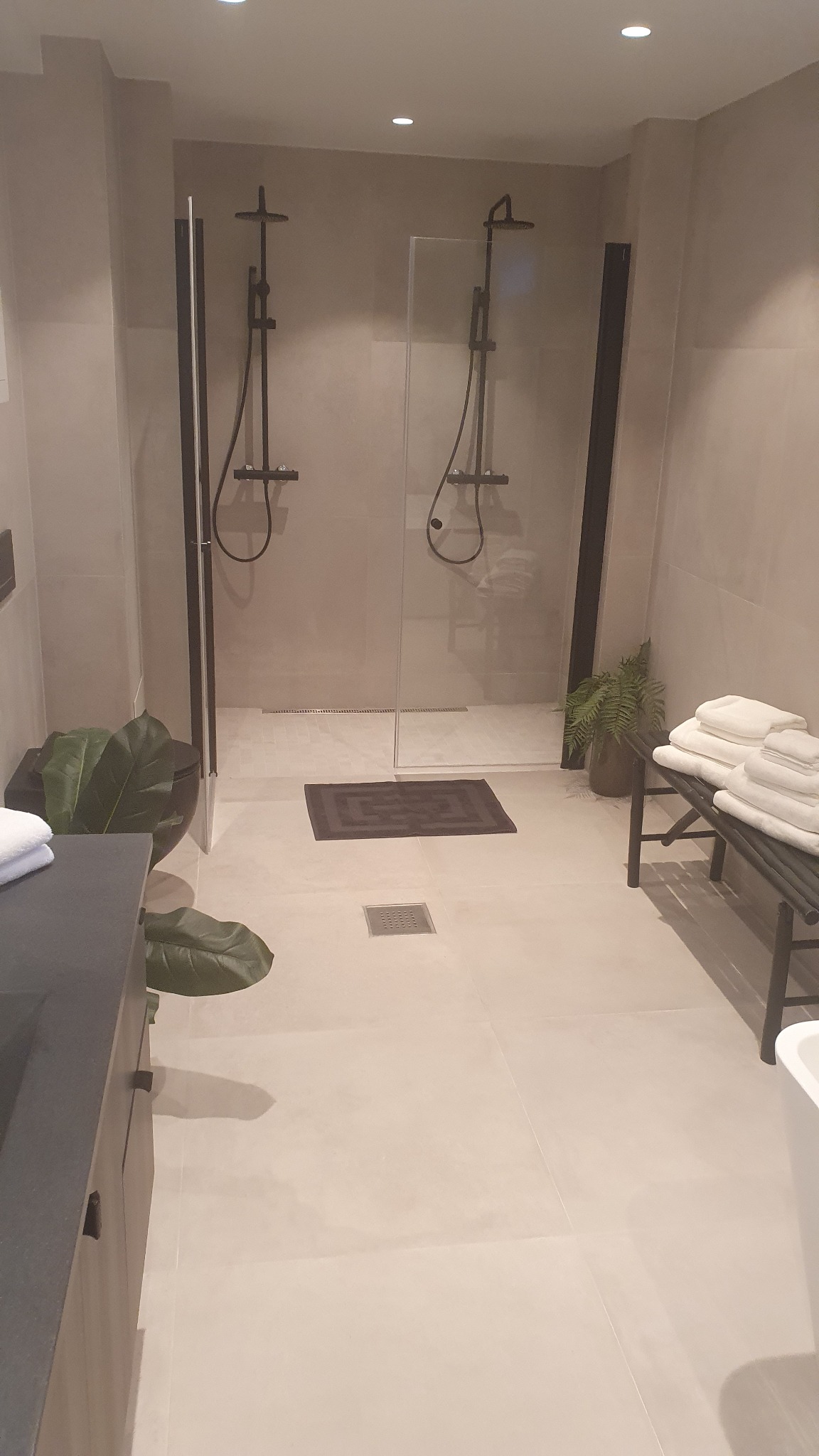 Minimalistyczna łazienka z dwoma kabinami prysznicowymi typu walk-in, beżowe płytki podłogowe i ścienne, czarne armatury, rośliny doniczkowe i sterta białych ręczników.