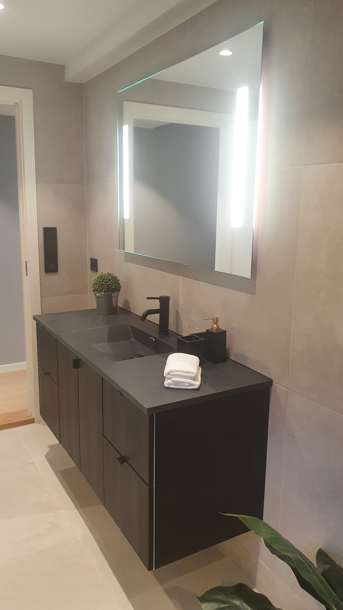 Nowoczesna łazienka z podświetlanym lustrem, ciemną szafką pod umywalką i beżowymi płytkami. Minimalistyczny design z rośliną doniczkową i białymi ręcznikami.