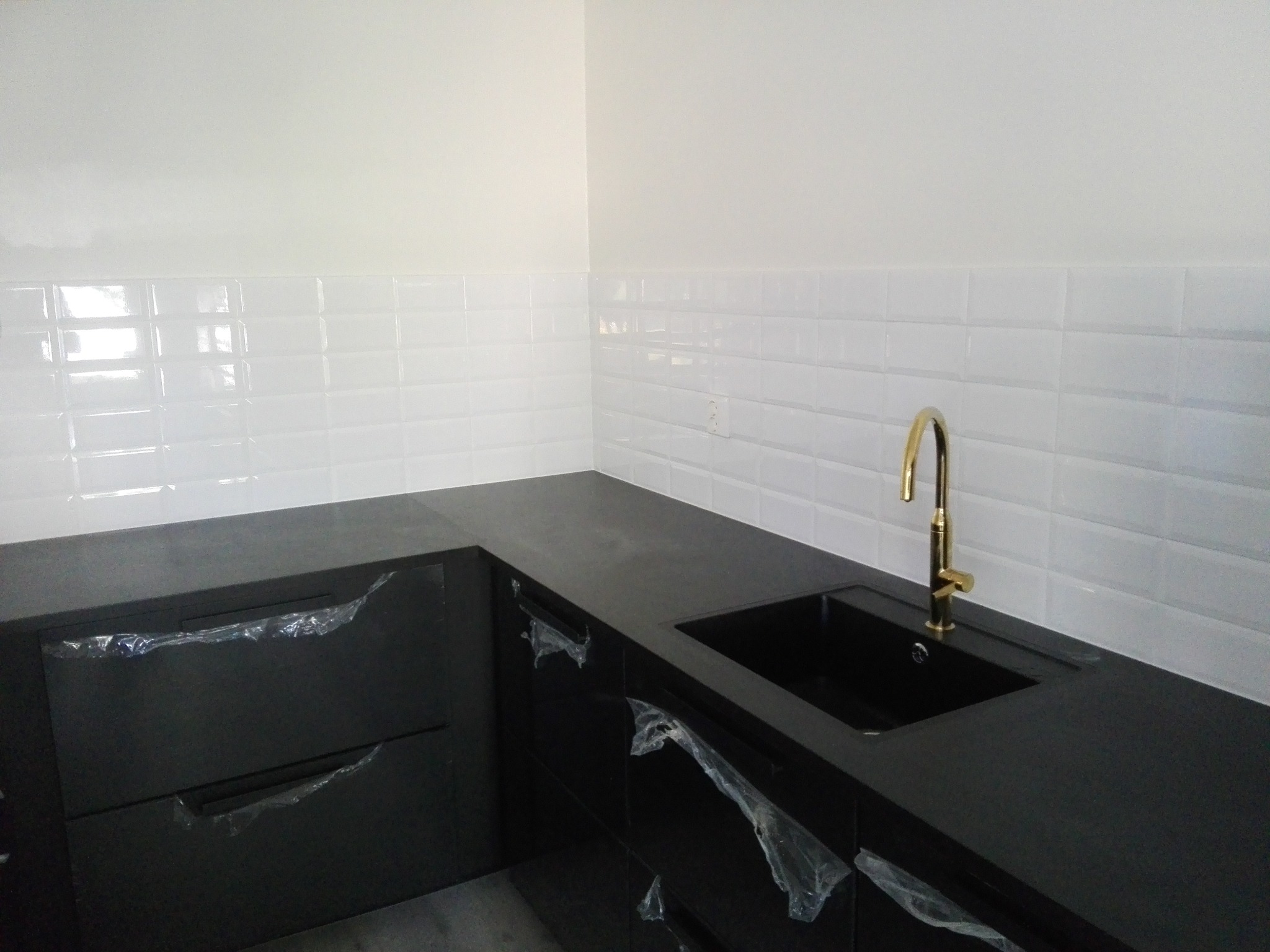 Nowoczesna, czarna zabudowa kuchenna z złotą baterią i białymi, błyszczącymi płytkami na ścianie. Elementy zabezpieczone folią ochronną.