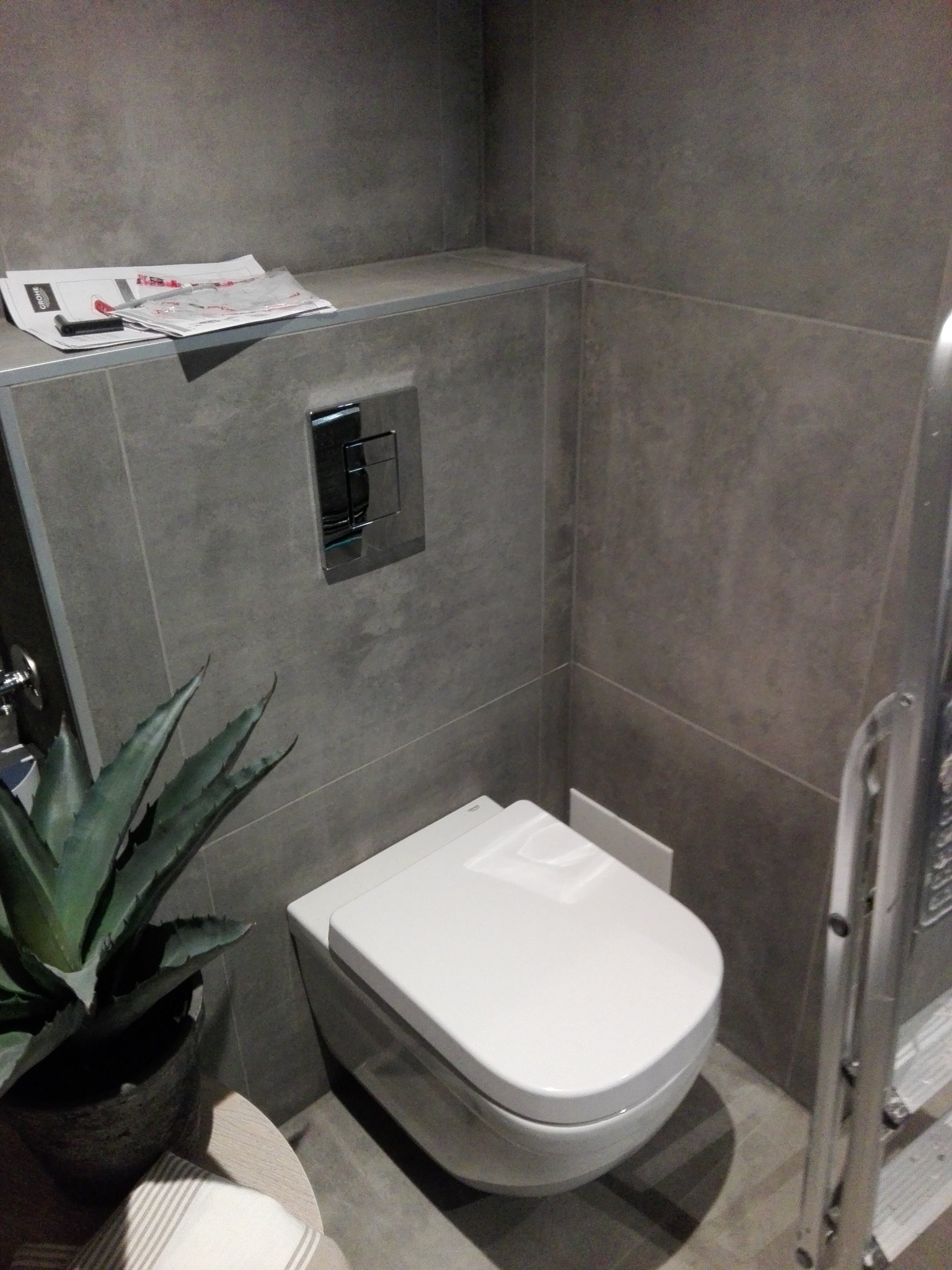 Nowoczesna toaleta podwieszana w szarej łazience, z widoczną instalacją podtynkową i rośliną doniczkową. Minimalistyczny design, płytki w odcieniach szarości.