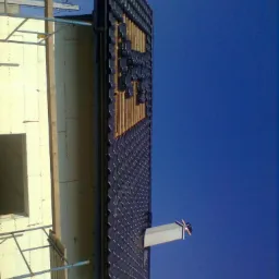 Montaż ciemnych dachówek na konstrukcji dachu, widoczne drewniane elementy i fragment elewacji budynku na tle intensywnego, błękitnego nieba.