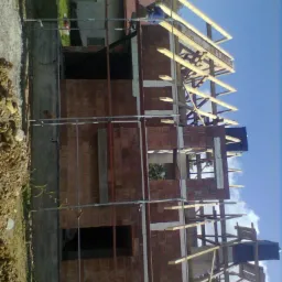 Budowa domu z cegły: wznoszenie ścian i konstrukcji drewnianej dachu, widoczne rusztowania i pracownik w niebieskim kombinezonie. Perspektywa z dołu, błękitne niebo.