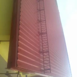 Czerwona blacha trapezowa na elewacji budynku z przymocowaną drabinką. Widok z dołu, ukośny kadr, fragment ściany i nieba w tle.