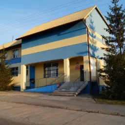 Dwukondygnacyjny budynek użytkowy z żółto-niebieską elewacją, jasnym dachem i schodami wejściowymi z metalową barierką. Widoczne drzewa iglaste i słup elektryczny.