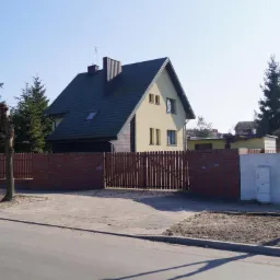 Żółty dom jednorodzinny z ciemnym dachem i drewnianą elewacją, otoczony ceglanym i drewnianym ogrodzeniem, widok z ulicy w słoneczny dzień.