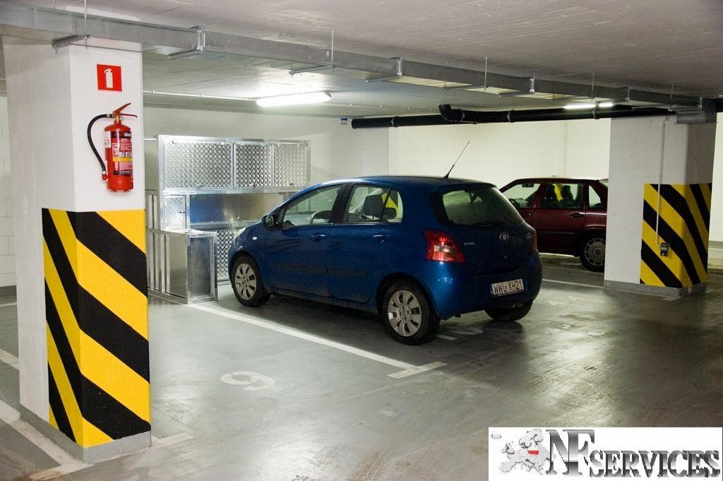 Aluminiowa zabudowa miejsc parkingowych w garażu podziemnym, widoczny niebieski samochód osobowy i słup z oznaczeniami ostrzegawczymi.