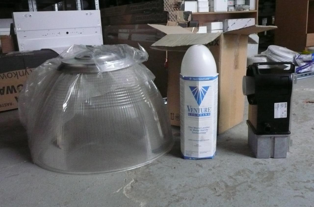 Klosz lampy przemysłowej w folii, obok opakowanie żarówki Venture Lighting i transformator, na tle kartonów i regałów magazynowych.