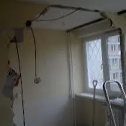 Pok&oacute;j w trakcie wyburzania ściany działowej, widoczne odsłonięte przewody elektryczne na suficie, fragmenty tapety na ścianie, okno z widokiem na budynek mieszkalny, drabina oparta o ścianę.