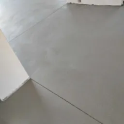 Świeżo wylana, gładka, szara podłoga z widocznymi dylatacjami, fragment ściany z wnęką w tle.