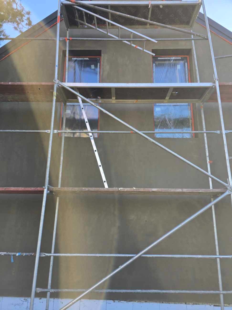 Ocieplony budynek w trakcie prac wykończeniowych. Widoczne rusztowania, okna zabezpieczone folią. Ściana pokryta szarym tynkiem. Prace na wysokości.