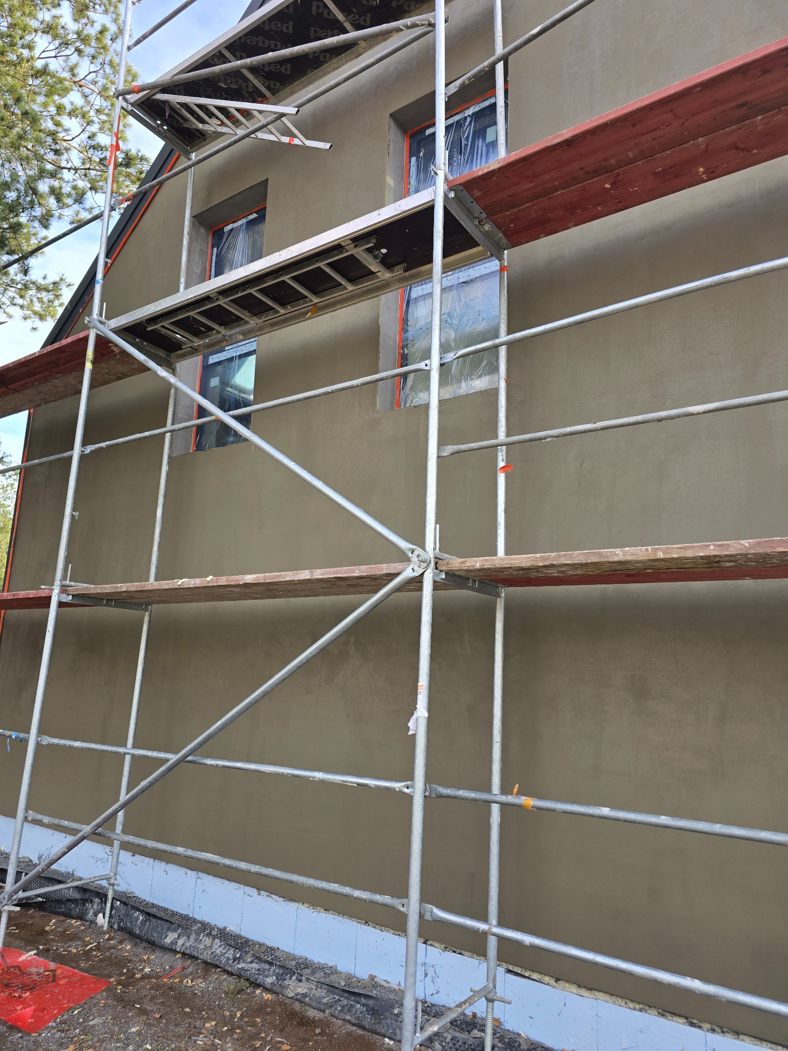 Elewacja domu w trakcie prac wykończeniowych, widoczne rusztowanie i okna zabezpieczone folią. Świeżo nałożony tynk w odcieniach szarości.