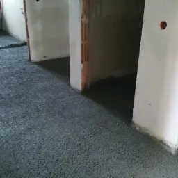 Świeżo wylana, szara posadzka w surowym stanie, widoczne niedoskonałości i nierówności przy ścianach, zarys przejść do kolejnych pomieszczeń z surowymi framugami.