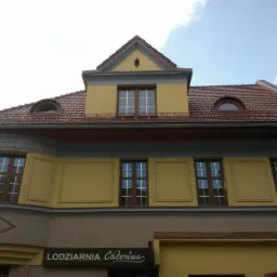 Budynek lodziarni Caterina z widocznym dachem z brązowej dachówki ceramicznej i żółtą elewacją w słoneczny dzień.