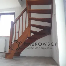 Drewniane schody ażurowe z balustradą, widok od dołu, z logo firmy stolarskiej na ścianie w tle.