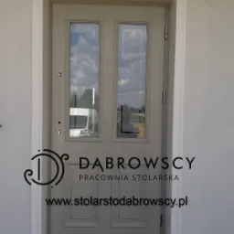 Szare drzwi wejściowe z dwoma szybami i białymi ramami, widoczne logo firmy stolarskiej i odbicie samochodu w szybach.