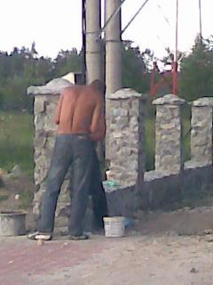 Mężczyzna bez koszulki, w jeansach, w trakcie murowania słupka ogrodzeniowego z kamienia. Widoczne wiadro z zaprawą i elementy ogrodzenia w tle.