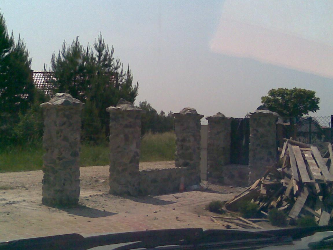 Kamienne słupki ogrodzeniowe z nakrywami w trakcie budowy, widoczny stos desek i niezabezpieczony teren.