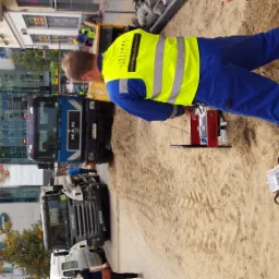 Pracownik w kamizelce, wykonujący badania gruntu w terenie miejskim przy użyciu specjalistycznego sprzętu. Na drugim planie budynki i samochody.