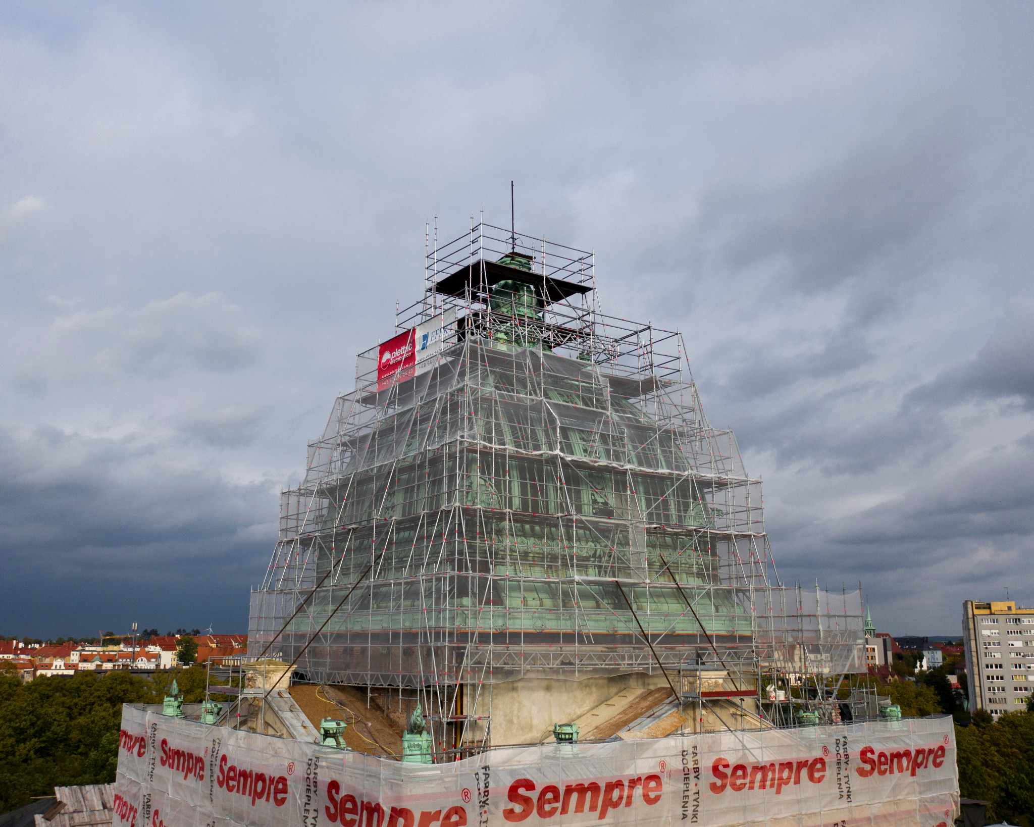 Kopuła budynku w rusztowaniach, osłonięta siatką z czerwonym logo 'Sempre', na tle pochmurnego nieba i zabudowy miejskiej. Prace na wysokościach.
