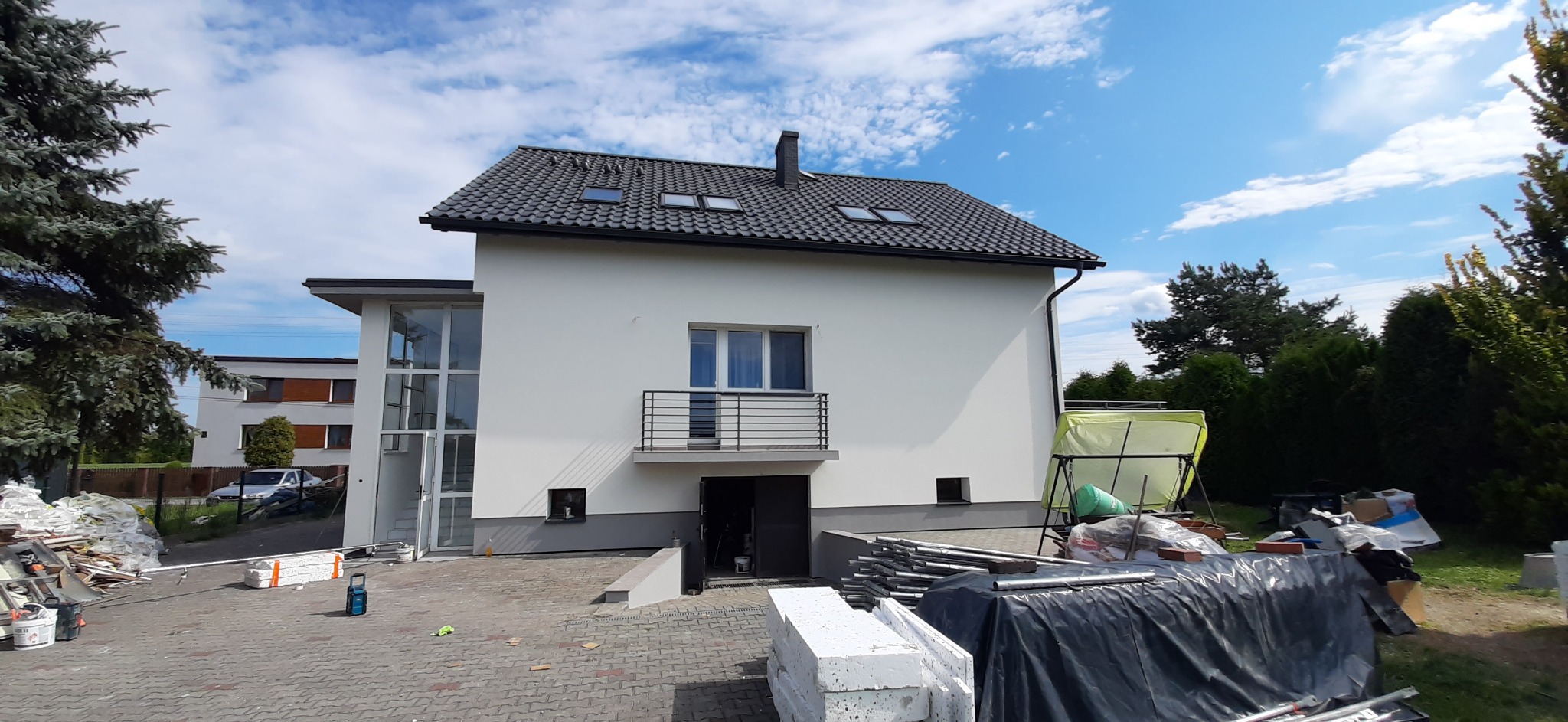 Biały, dwukondygnacyjny dom z szarą dachówką i balkonem, po świeżym malowaniu elewacji, widoczne materiały budowlane na posesji.