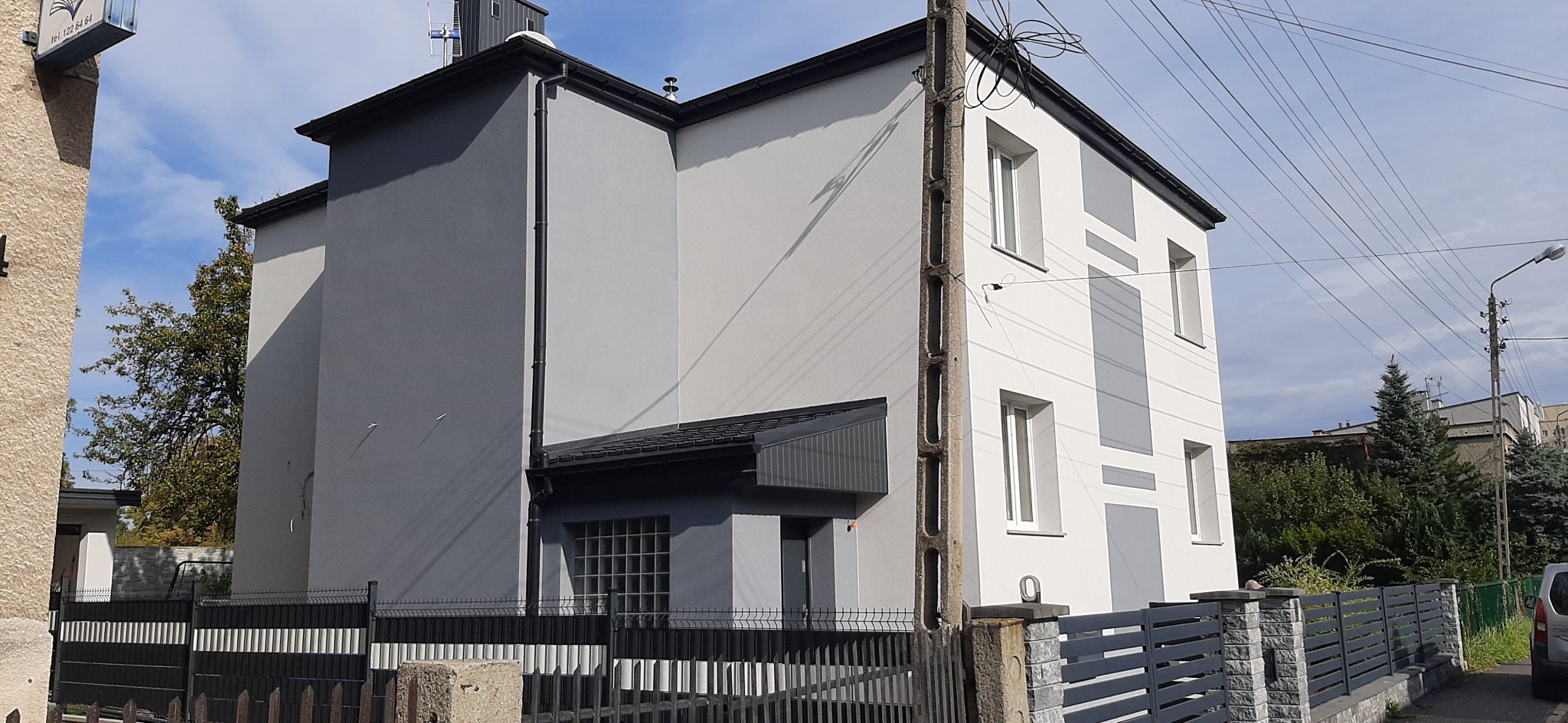 Dwukondygnacyjny budynek po remoncie elewacji, z szarą i białą kolorystyką, widoczny zza metalowego ogrodzenia, z słupem elektrycznym na pierwszym planie.