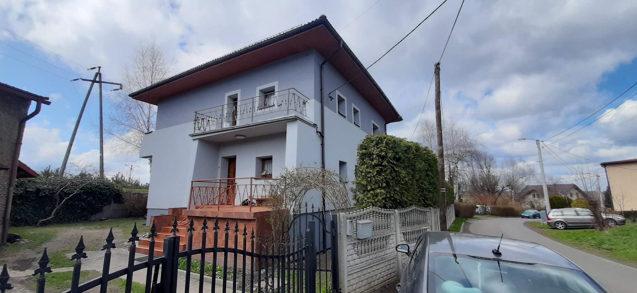 Dwupiętrowy dom z balkonem, szarą elewacją i brązowym dachem, widoczny zza czarnego, kutego ogrodzenia.