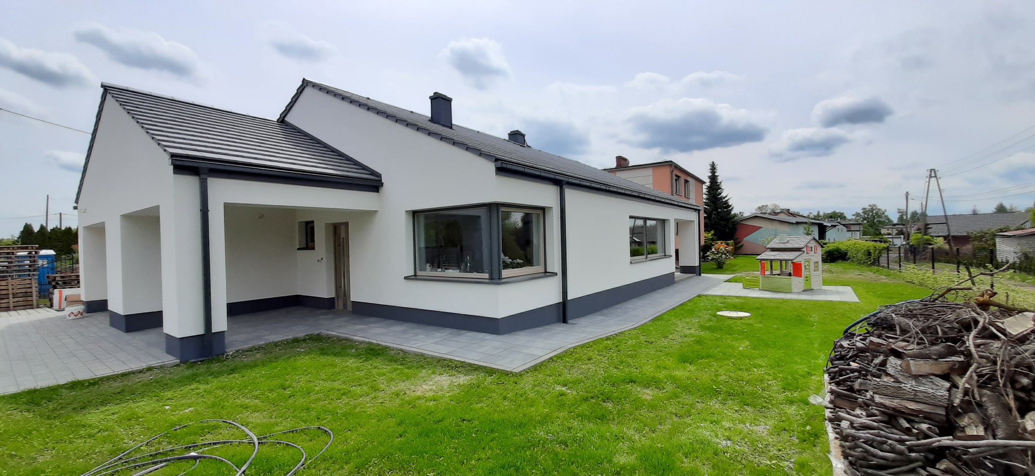 Nowoczesny dom jednorodzinny z białą elewacją i szarą podmurówką, widok z zewnątrz, z zadbanym trawnikiem i stertą drewna w rogu kadru.