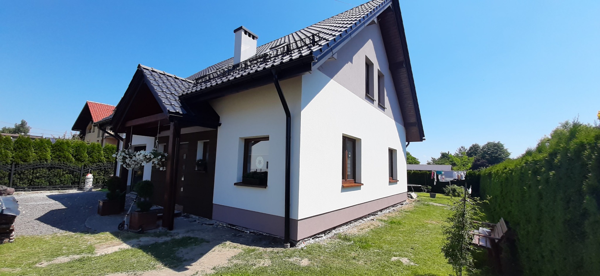 Nowoczesny dom jednorodzinny z białą elewacją, ciemną podmurówką i dachem z czarnej dachówki, widok z boku na tle błękitnego nieba i zielonego ogrodu.