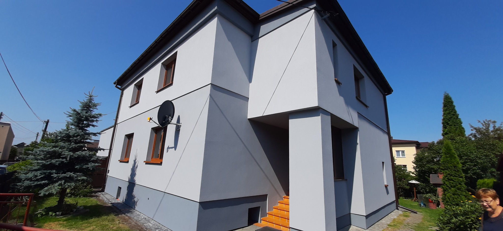 Dwukondygnacyjny dom jednorodzinny z nową, dwukolorową elewacją w odcieniach szarości, widoczna antena satelitarna, pomarańczowe schody wejściowe i iglaki w ogrodzie.