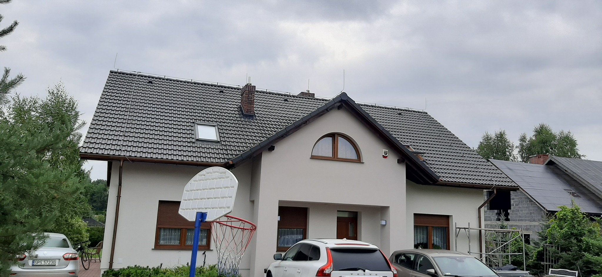 Dwupiętrowy dom jednorodzinny z jasną elewacją, ciemną dachówką i brązowymi ramami okiennymi, z przodu widoczny kosz do koszykówki i zaparkowane samochody.