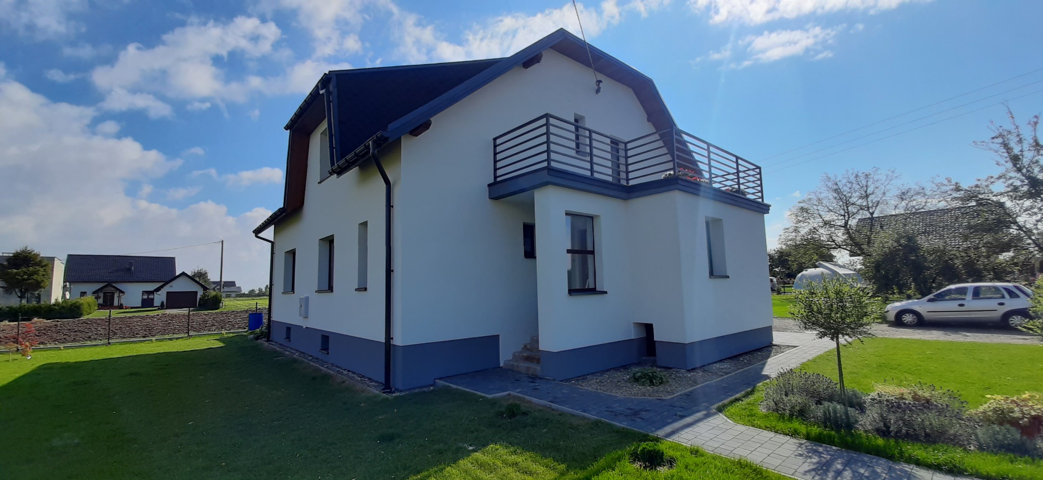 Nowoczesny dom jednorodzinny z białą elewacją, ciemnoszarą podmurówką i balkonem z metalową balustradą, otoczony zielonym trawnikiem i kostką brukową, widok z boku w słoneczny dzień.