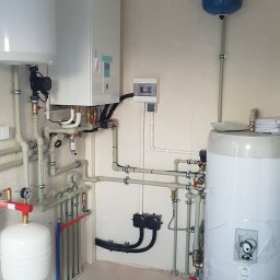 EKOWENT INSTAL - Złożona instalacja grzewcza w kotłowni: biały zasobnik ciepłej wody, ekspansomat, rozdzielnica elektryczna i rury z zaworami. Widoczne elementy instalacji rekuperacji.