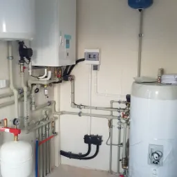 Złożona instalacja grzewcza w kotłowni: biały zasobnik ciepłej wody, ekspansomat, rozdzielnica elektryczna i rury z zaworami. Widoczne elementy instalacji rekuperacji.