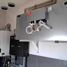 EKOWENT INSTAL - Szara centrala wentylacyjna z podłączonymi przewodami, zamontowana na ścianie w pomieszczeniu technicznym obok kotła grzewczego i instalacji rurowej.