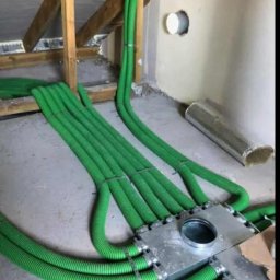 EKOWENT INSTAL - Zielone rury wentylacyjne przymocowane do metalowej skrzynki rozdzielczej w surowym wnętrzu poddasza, widoczne drewniane elementy konstrukcyjne.