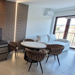 EKOWENT INSTAL - Nowoczesne wnętrze z klimatyzatorem ściennym: minimalistyczny design, drewniane panele z oświetleniem, okrągły stół z krzesłami i biała sofa przy oknie.