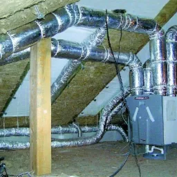 Szara jednostka centralna z podłączonymi, izolowanymi przewodami wentylacyjnymi w przestrzeni poddasza z widoczną izolacją i drewnianą konstrukcją dachu.