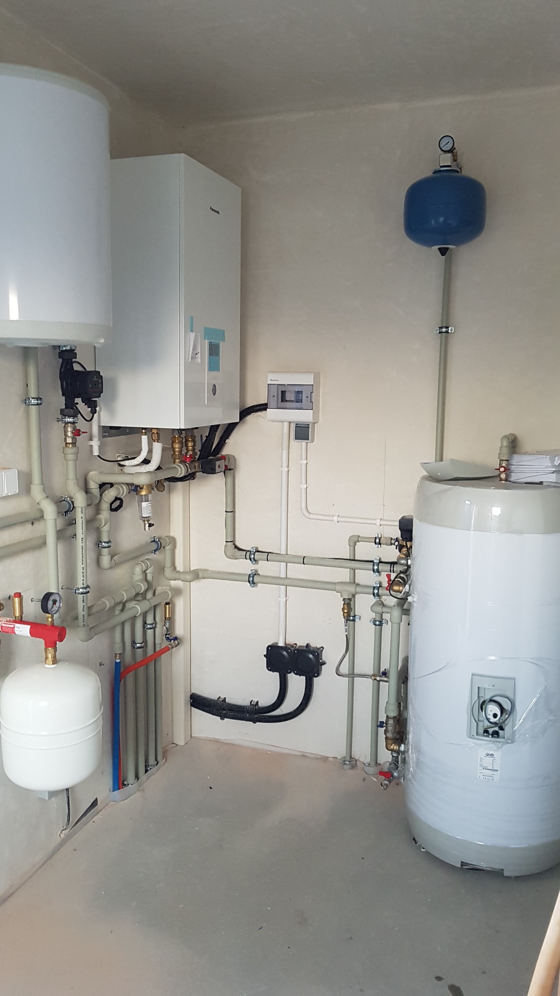 Złożona instalacja grzewcza w kotłowni: biały zasobnik ciepłej wody, ekspansomat, rozdzielnica elektryczna i rury z zaworami. Widoczne elementy instalacji rekuperacji.