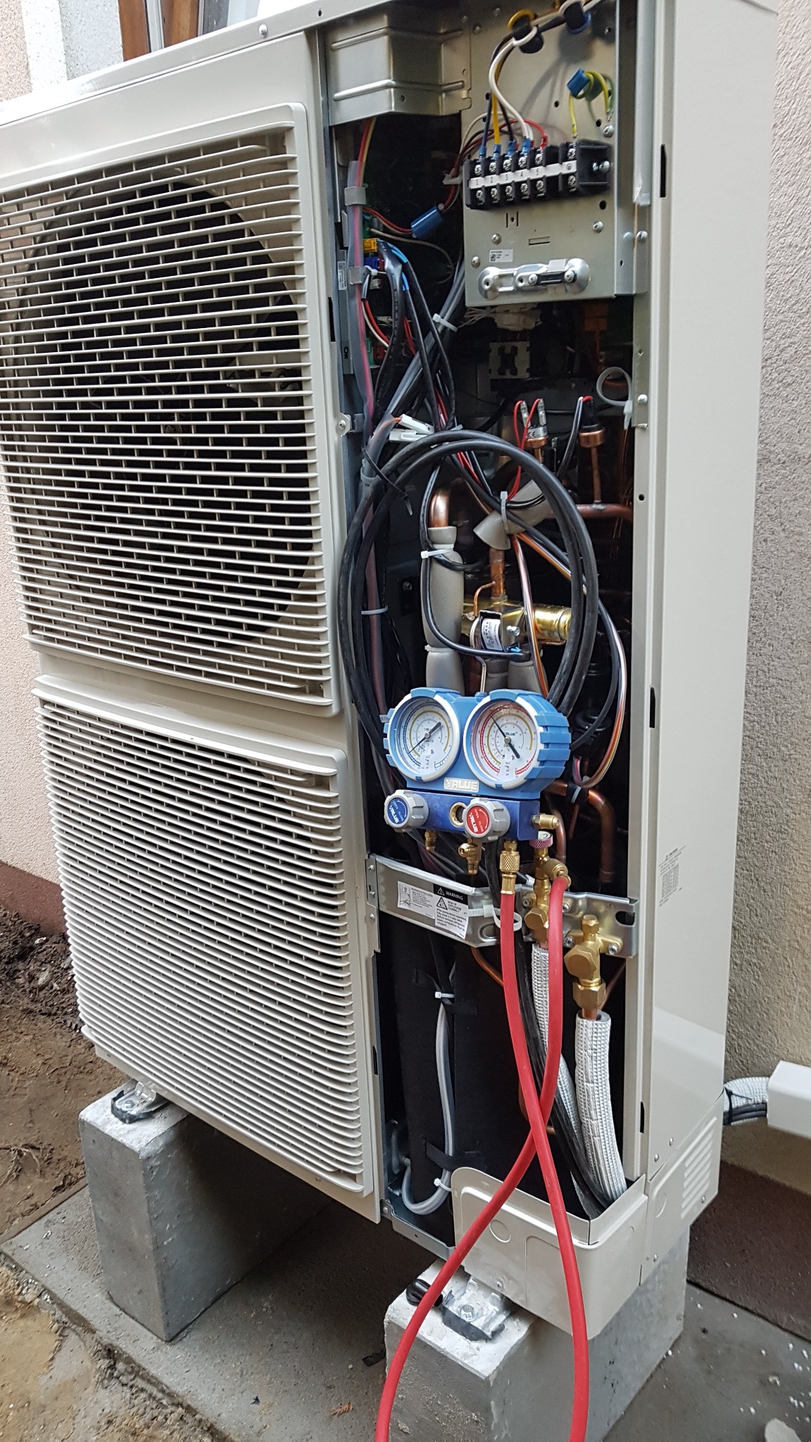 Otwarta obudowa klimatyzatora z widocznymi przewodami, manometrami i elementami instalacji, umieszczona na betonowych blokach przy ścianie budynku.