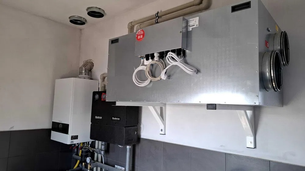 Szara centrala wentylacyjna z podłączonymi przewodami, zamontowana na ścianie w pomieszczeniu technicznym obok kotła grzewczego i instalacji rurowej.