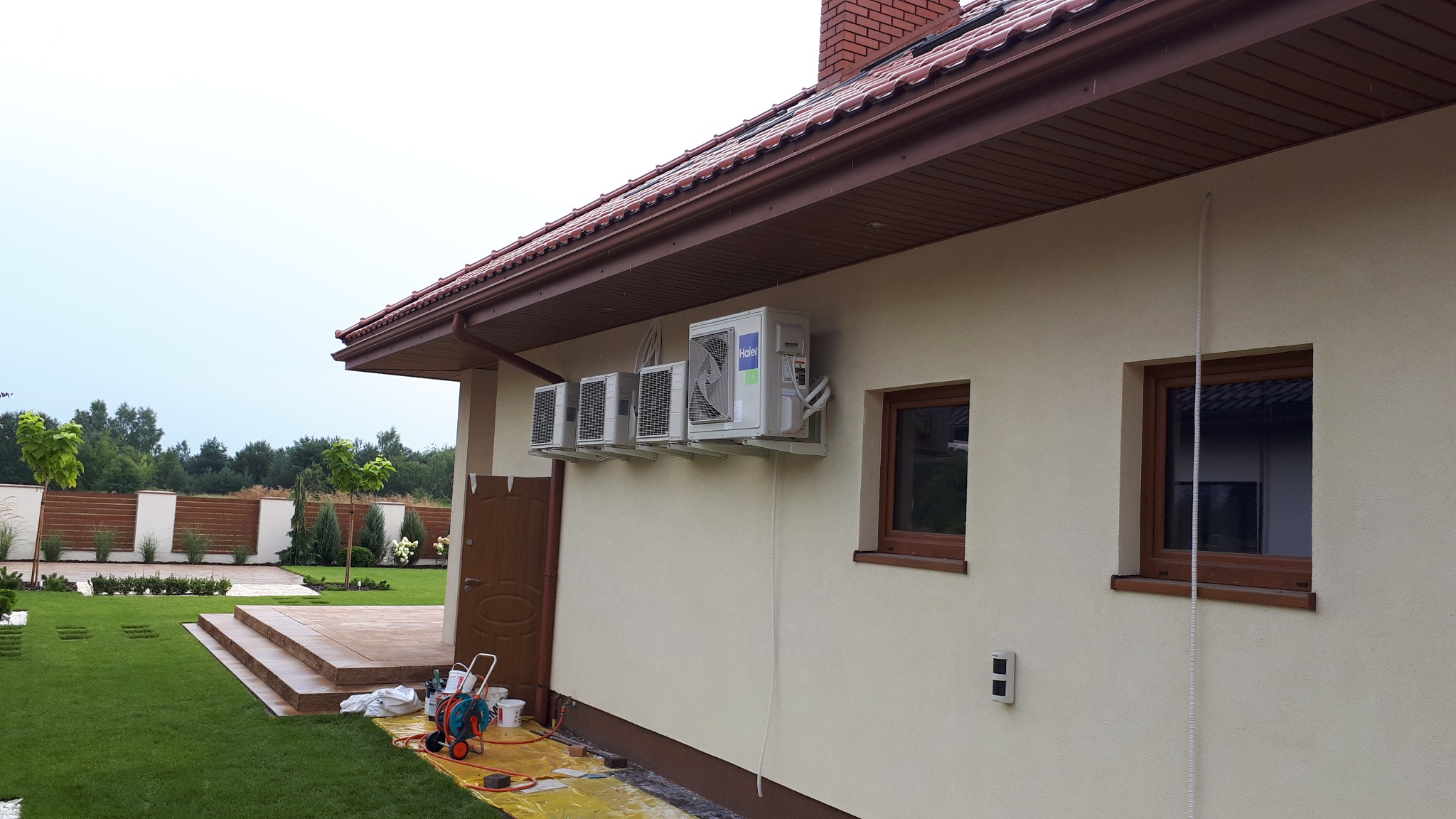 Cztery klimatyzatory Haier zamontowane na elewacji domu jednorodzinnego, widoczne narzędzia i materiały montażowe, ogród w tle.
