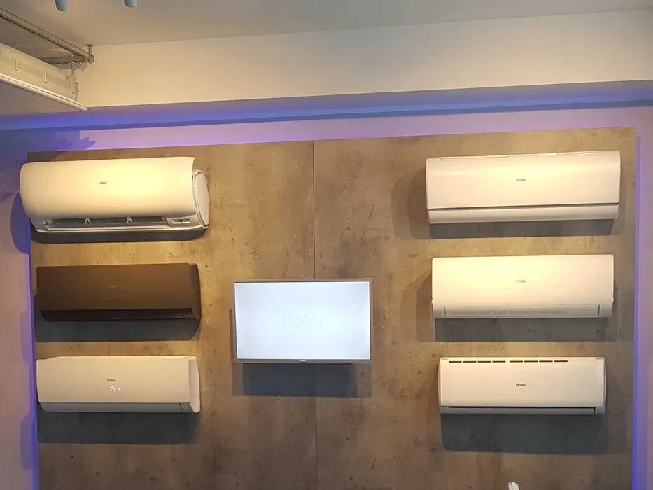 Ekspozycja klimatyzatorów Haier na ścianie o teksturze betonu. Różne modele i kolory, w tym białe i brązowe. Centralnie umieszczony ekran z logo Haier.