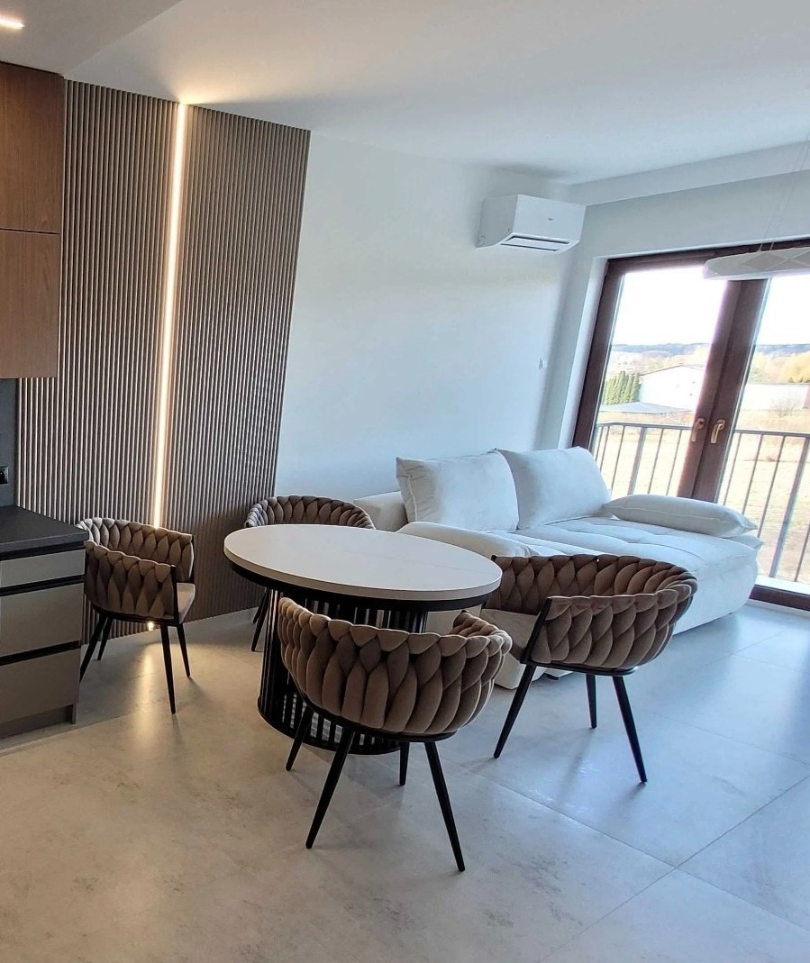 Nowoczesne wnętrze z klimatyzatorem ściennym: minimalistyczny design, drewniane panele z oświetleniem, okrągły stół z krzesłami i biała sofa przy oknie.