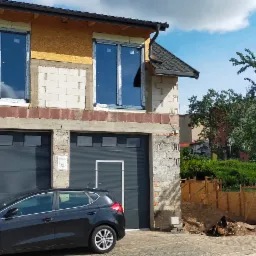 Dom w trakcie budowy z zamontowanymi oknami na piętrze i dwiema szarymi bramami garażowymi na parterze, obok zaparkowany czarny samochód.