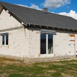 Surowy stan domu jednorodzinnego z zamontowanymi oknami, częściowo z roletami zewnętrznymi, białe ściany z betonu komórkowego, szara dachówka, widoczne prace wykończeniowe.