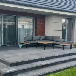 Nowoczesny taras z szarych płyt z niskimi schodami, wyposażony w sofę z szarymi poduszkami i stoliki kawowe, z przesuwnymi drzwiami na taras z siatką przeciw owadom i widokiem na ogród z różowym...