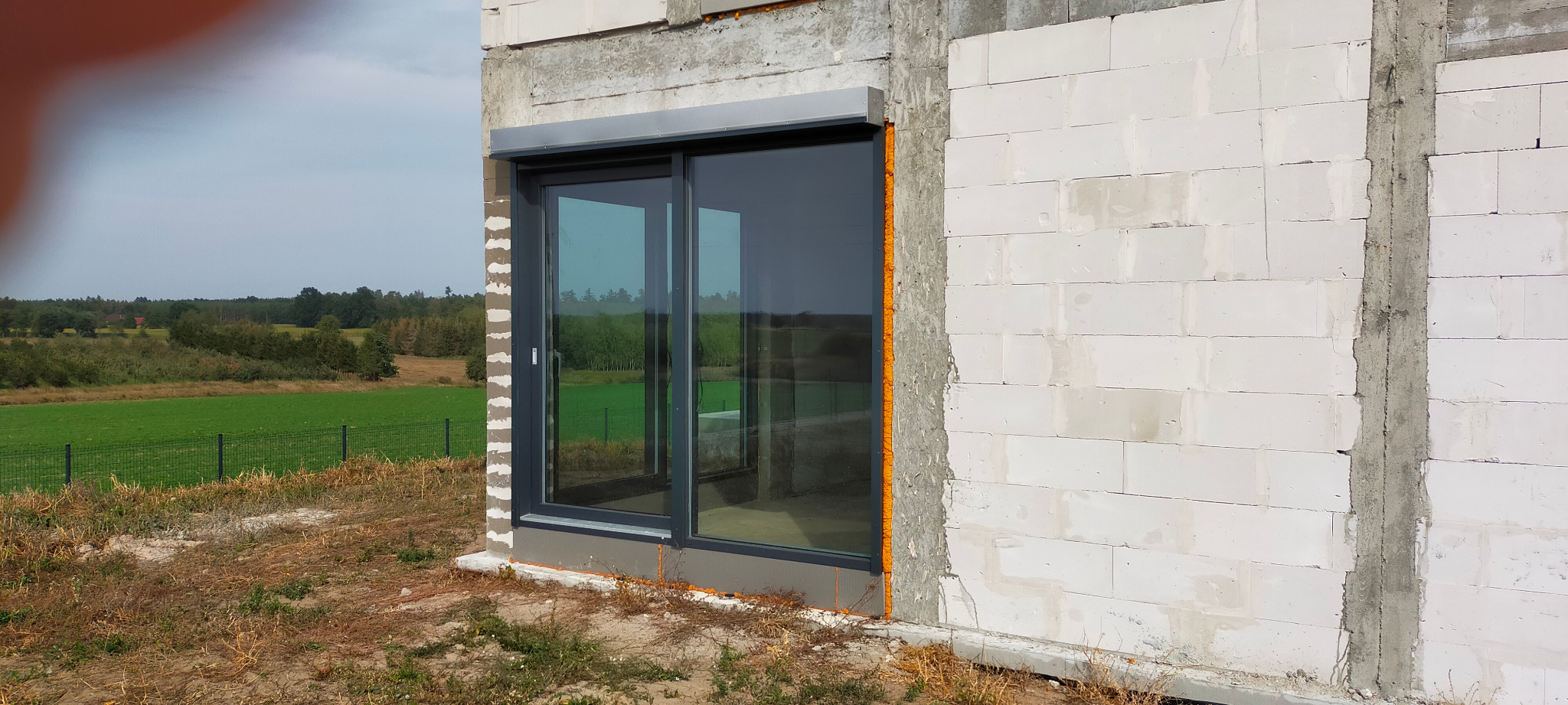 Montaż okna przesuwnego w surowym stanie budynku z bloczków, widoczne pianowanie wokół ramy i krajobraz za oknem.