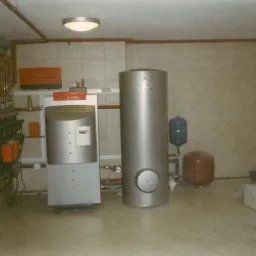 Kotłownia z zainstalowanym piecem i zasobnikiem wody, widoczne rury grzewcze, manometry i zbiorniki wyrównawcze w pomieszczeniu z wykończonymi ścianami i podłogą.