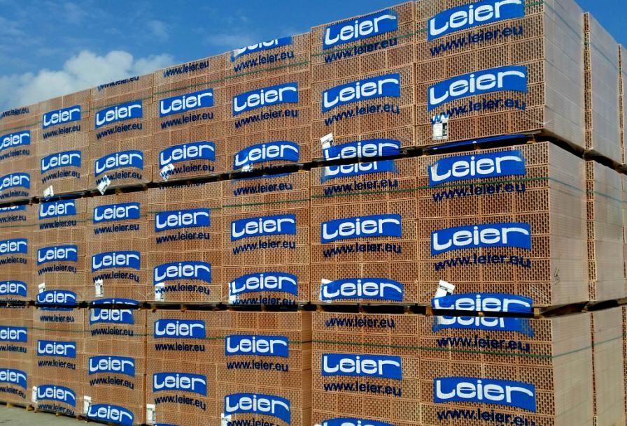Palety z pustaków ceramicznych Leier, zabezpieczone folią i taśmami, składowane na placu budowy pod błękitnym niebem.