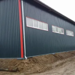 Nowoczesna hala z płyt warstwowych w kolorze grafitowym, z czerwonymi akcentami i białymi oknami, widoczna od strony elewacji bocznej, w tle drzewa.